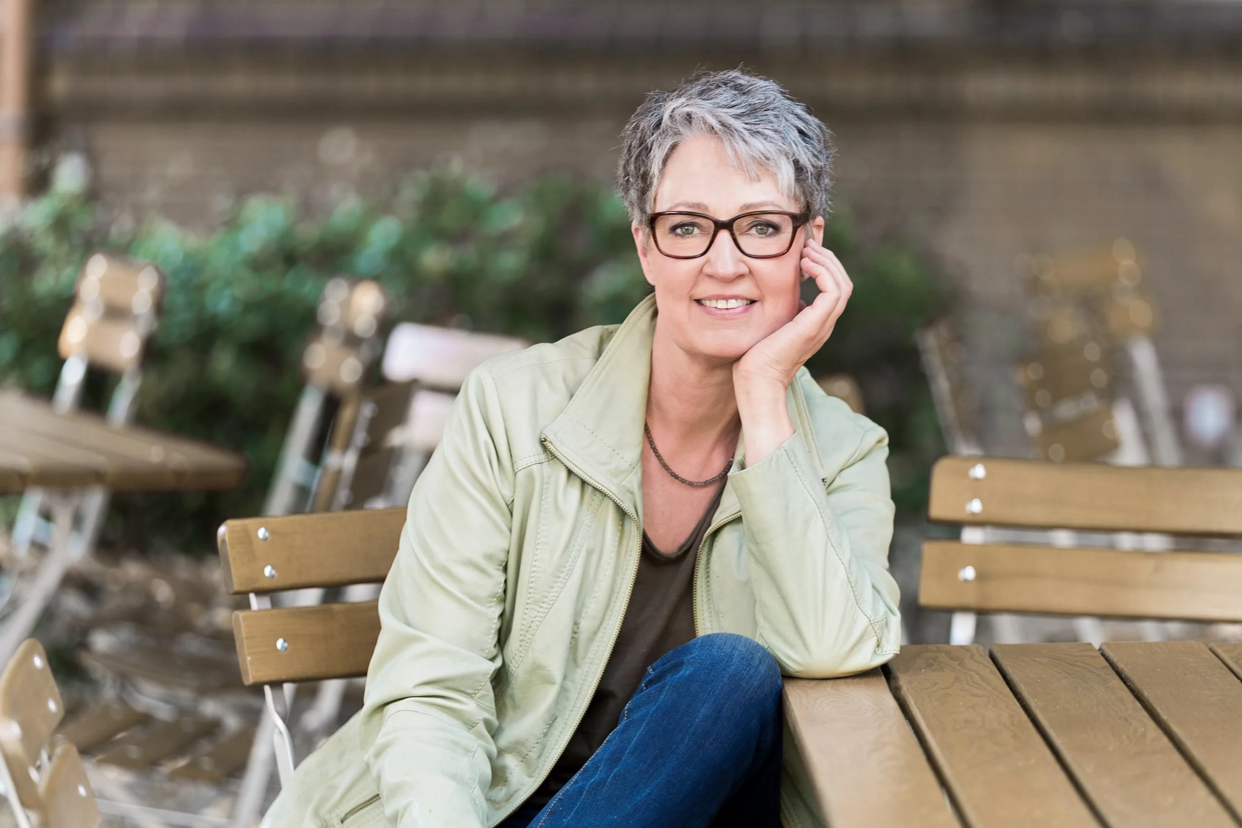 Portraitfoto von Vocalcoach Birgitta Schaub, an einem Tisch auf einer Terrasse freundlich lächelnd